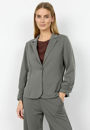 Egyenes szőke hajú nő szürke blézerben és hozzá illő nadrágban, bordó felsővel, egyik kezét a zsebében tartva áll.