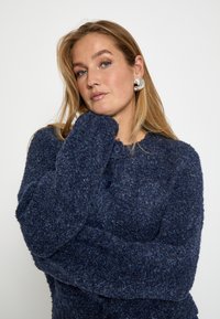 Jūras zila teksturēta cardigan jakas ar apaļu kakla izgriezumu, garām piedurknēm un lielām pogām. Matu stils nolaižams, rotāts ar sudraba izteiksmīgām auskarām.