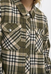 Chemise en flanelle à carreaux verts et noirs avec des accents orange et blanc, dotée d'un col pointu, de deux poches poitrine et de boutons-pression en métal.