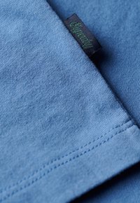 Tessuto di cotone azzurro chiaro con una texture morbida, caratterizzato da un bordo ripiegato e un'etichetta nera con testo verde. Dettaglio ravvicinato della cucitura.