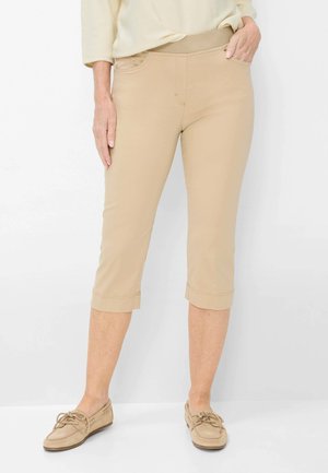 Kvinde iført beige kortere bukser med opkørte kanter og matchende loafers, stående foran en hvid baggrund.