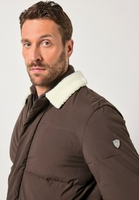 Man met kort bruin haar en baard draagt een bruine gewatteerde jas met een witte fleece kraag, staand tegen een effen lichte achtergrond.
