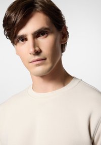 Jasno beżowy sweterek z okrągłym dekoltem, wykonany z miękkiego materiału, wyróżniający się gładką fakturą i prostym wzornictwem, bez widocznych wzorów czy akcentów.