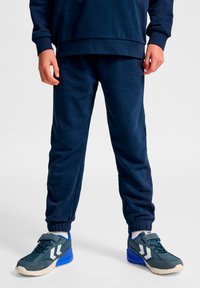 Granatowe spodnie dresowe z elastycznymi ściągaczami i bocznymi kieszeniami, połączone z ciemnozielonymi sneakersami z białymi akcentami i niebieskimi detalami.