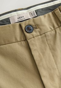 Khaki nohavice s voľným strihom, s gombíkovým zapínaním, zipsom a kontrastnými pásikmi na páse. Vyrobené z odolného materiálu s hladkou textúrou.