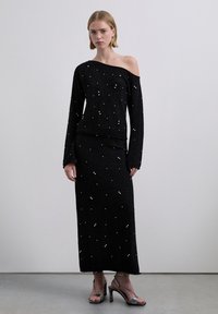 Conjunto de dos piezas de punto negro que incluye un top de manga larga tipo off-shoulder y una falda maxi, adornados con pequeñas decoraciones que parecen perlas.