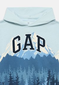 Jasnoniebieska bluza z kapturem z logo "GAP" w ciemnoniebieskim kolorze, przedstawiająca grafikę gór i lasu w różnych odcieniach niebieskiego i zielonego. Materiał bawełniany.