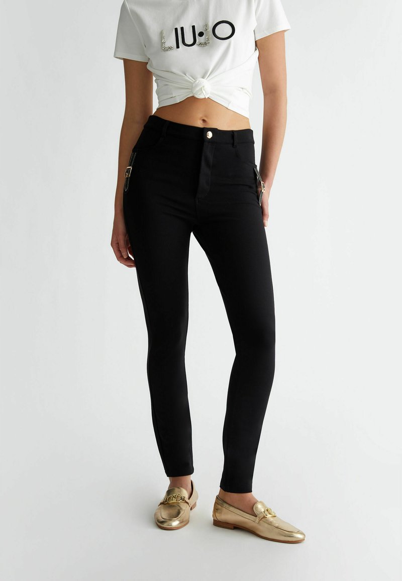 Melnas pieguļošas bikses ar sānu rāvējslēdzējiem un spīdīgu zelta pogu, apvienotas ar mezglošu baltu T-kreklu un metāliskām mokasīnēm.