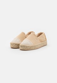 Beige espadriller med et teksturert lerretsoverdel, frynsete kanter og en vevd jutesåle. Rund tå med kontrasterende lyse detaljer.