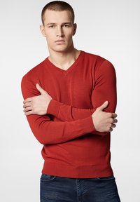 Czerwony sweter dzianinowy z dekoltem w serek, długimi rękawami oraz prążkowanymi mankietami i dołem, noszony na niebieskich podartych dżinsach. Gładka tekstura, dopasowany krój.