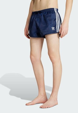 Personne aux jambes nues portant un short Adidas bleu marine avec des bandes blanches sur les côtés, debout pieds nus sur un fond clair.