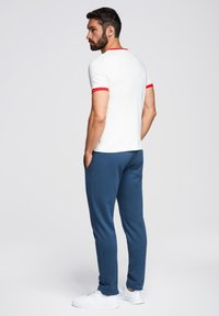 Vit t-shirt med röda detaljer på ärmarna och kragen, ihop med marinblå joggers. Modellen vänder sig bort, har händerna i fickorna och bär vita sneakers.