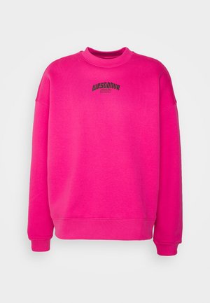Osoba ubrana w jaskraworóżową bluzę, czarne spodnie, beżowo-pomarańczowe sneakersy oraz różowo-czarną saszetkę The North Face, stojąca na tle szarej ściany.