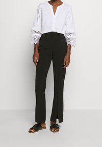 Camisa blanca con mangas de campana rematadas en encaje, combinada con pantalones negros de cintura alta que tienen aberturas laterales y sandalias negras de tiras.