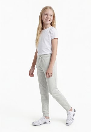 Nuori tyttö, jolla on pitkät vaaleat hiukset, pukeutuneena valkoiseen t-paitaan, vaaleanharmaisiin collegehousuihin ja valkoisiin lenkkareihin, seisoo ja katsoo sivulle.