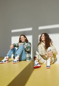 Deux jeunes adultes assis sur un sol jaune contre un mur, souriant, vêtus de vêtements décontractés et portant des baskets blanches, rouges et noires assorties.