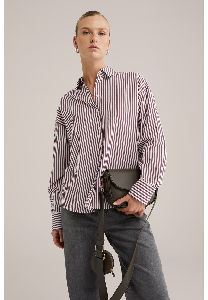 Camicia a righe rossa e bianca, abbinata a jeans grigi. Tiene una borsa a tracolla verde scuro con un accento circolare.