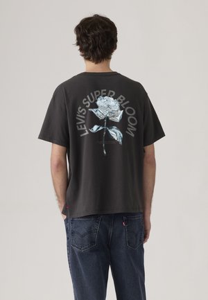 Homme aux cheveux bouclés foncés portant un t-shirt noir avec une rose argentée métallique et le texte « Levi's Super Bloom » dans le dos, associé à un jean bleu foncé.