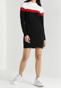 Zwart gebreid jurk met witte en rode horizontale strepen op de borst, met een hoge hals en lange mouwen, gecombineerd met witte sneakers.