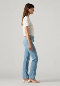 Jeans acampanados de color azul claro con cintura alta, bolsillos traseros con costuras en forma de corazón, combinados con una camiseta blanca de manga corta y sandalias.