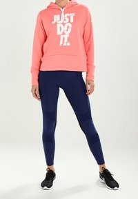 Koraal hoodie met witte "JUST DO IT." tekst, gecombineerd met navy leggings met textuurpanelen, en zwarte sport schoenen.
