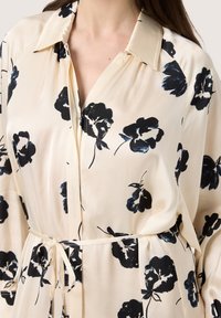 Bluză bej deschis cu imprimeu floral negru, cu guler, nasturi la față, legătură la talie și textură moale de satin. Mâneci lungi cu croială lejeră.