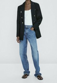 Blazer noir croisé à boutons dorés, débardeur blanc, jean bleu clair à jambes droites, et tongs noires. Design minimaliste.