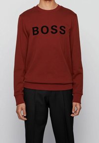 Sweatshirt bordeaux à manches longues, col rond côtelé et inscription noire « BOSS » sur la poitrine. Tissu lisse, coupe décontractée.