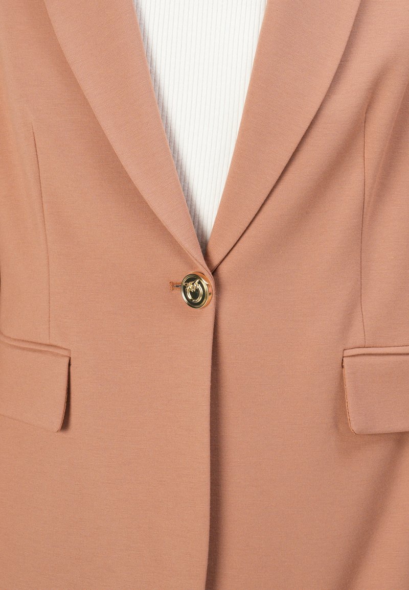 Różowa marynarka wykonana z gładkiego materiału, z pojedynczym zapięciem na złoty guzik i dwiema przednimi kieszeniami. Widoczna teksturowana biała podszewka.