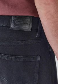 Lähedal vaade inimesest, kes kannab musti teksaseid nahkpatchiga, millel on kirjas "Denim Goods", ja sisenenud burdeos värvi särki.