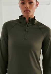Top sportiv de culoare verde închis, cu jumătate de fermoar, confecționat dintr-un material neted și ușor, prezentând un guler în picioare și mâneci lungi. Detalii cu cusături vizibile.