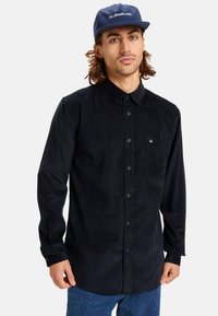 Black corduroy shirt with a button-up design, featuring a chest pocket, long sleeves, and a textured finish. Worn with a blue cap.