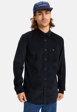 Black corduroy shirt with a button-up design, featuring a chest pocket, long sleeves, and a textured finish. Worn with a blue cap.