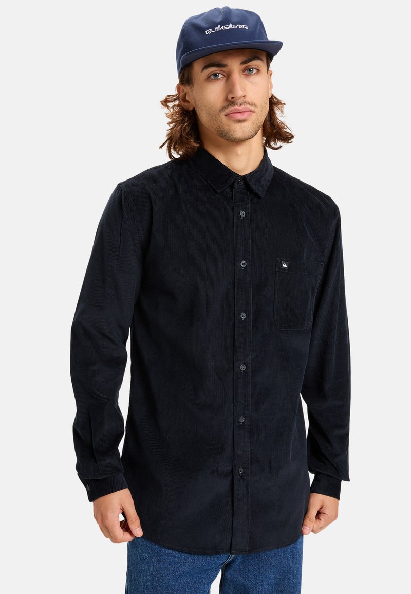 Black corduroy shirt with a button-up design, featuring a chest pocket, long sleeves, and a textured finish. Worn with a blue cap.