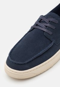 Zapato de ante azul marino con punta redondeada, costuras sutiles y suela de goma ligera en contraste; cuenta con cierre de cordones planos y un diseño minimalista.