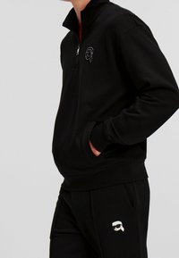 Sudadera negra con cremallera y cuello alto, puños de canalé y bolsillos laterales. Presenta un logo de dibujos animados blanco en el pecho. Textura de tela suave.