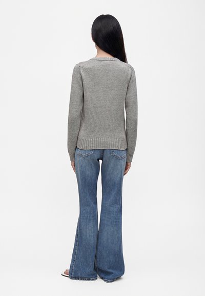 Femme aux longs cheveux foncés portant un pull en maille gris et un jean flare bleu, debout de dos sur un fond blanc.