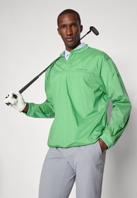 Lichtgroene lichtgewicht jas met een staande kraag, lange mouwen en elastische manchetten. Draagt een grijze broek en een golfclub. Glad textuur.