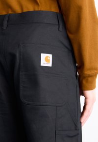 Černé cargo kalhoty s kapsou na zadní straně a bílou logovou nášivkou. Materiál vypadá odolně, s viditelným švem a hladkou texturou.