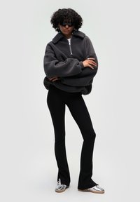 Szary polarowy sweter z zamkiem błyskawicznym i kołnierzem, zestawiony z czarnymi dzwonami i srebrnymi sneakersami. Modelka nosi eleganckie czarne okulary przeciwsłoneczne.