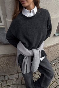 Szary sweter o luźnym kroju z prążkowanymi mankietami, noszony na białej koszuli z guzikami. Jasnoszary sweter zawiązany w talii. Ciemne spodnie.