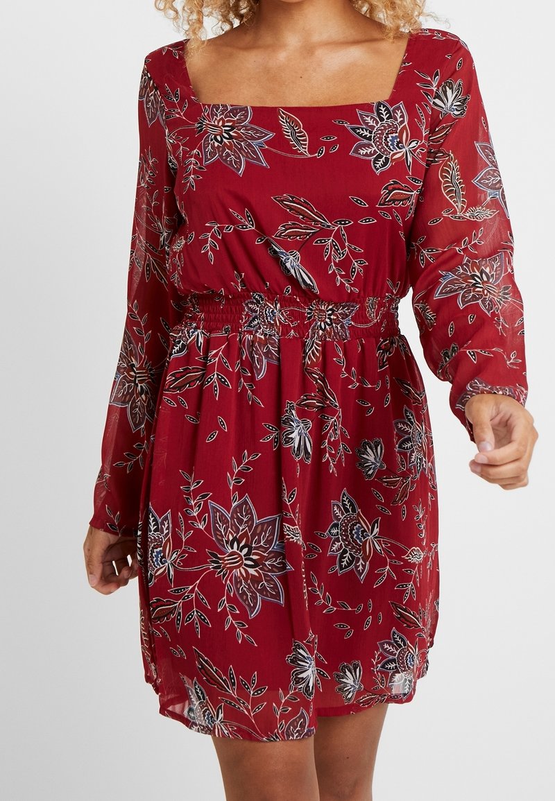 Femme portant une robe rouge à fleurs à manches longues avec un décolleté carré et une taille élastique, debout devant un fond uni.