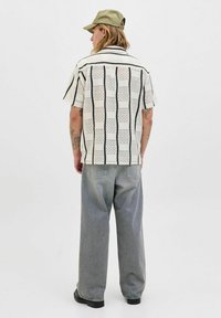 Chemise à manches courtes avec une base blanche, des rayures verticales noires et des motifs complexes. Associée à un jean gris ample et une casquette verte.