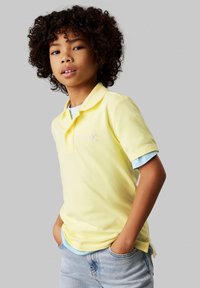 Polo di cotone giallo con colletto classico, caratterizzato da un piccolo logo ricamato, indossato sopra una maglietta azzurra chiara. Completato con jeans di denim chiaro.