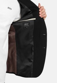 Hand hält offene schwarze maßgeschneiderte Jacke, die das glatte Futter und das innenliegende Etikett mit dem Markennamen JEFF und Größe 50 zeigt.