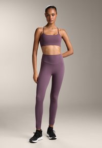 Vijolična športna srajčka in hlače z visoko pasom iz gladkega materiala, v kombinaciji s črnimi športnimi čevlji. Gladek tekstura in prilega obliko.