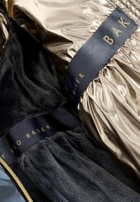 Metallisk guld yttertyg med en slät textur, fodrat med mjuk svart konstpäls. Har marinblå Ted Baker-logotyp.
