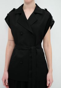 Gilet double boutonnage sans manche noir avec épaulettes, taille ceinturée et deux poches avant. Tissu texturé avec des accents de boutons.