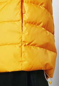 Veste matelassée jaune vif avec surpiqûres horizontales, dotée d'une fermeture éclair latérale et d'un cordon de serrage avec un bouton jaune à l'ourlet.