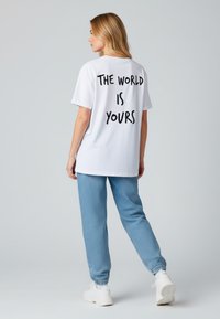 Witte T-shirt met zwarte tekst "DE WERELD IS VAN JOU" op de achterkant, gecombineerd met lichtblauwe joggers en witte sneakers. Glad materiaal, ontspannen pasvorm.